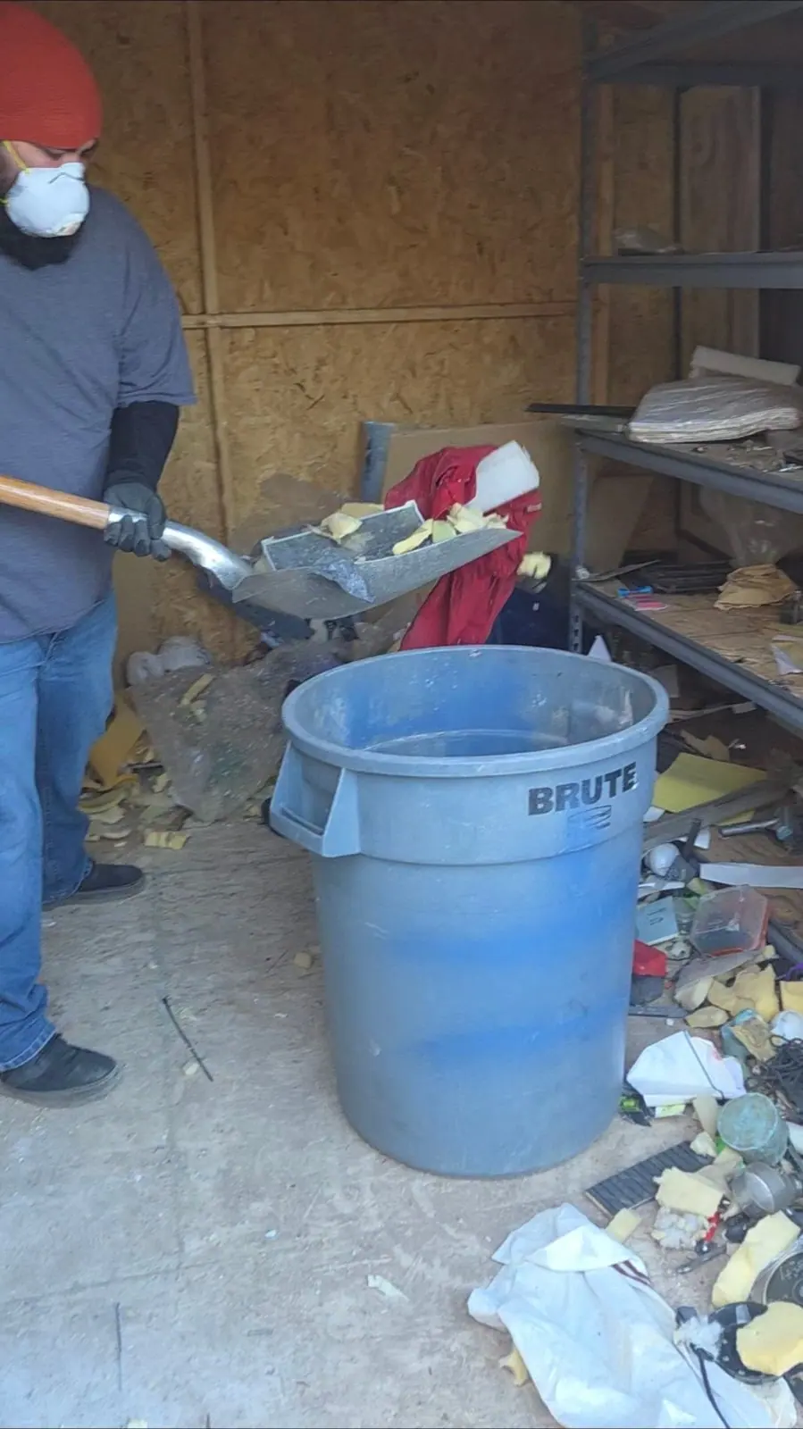 Henderson Junk Solutions crew member in a mask and gloves shoveling debris into a trash can inside a shed during a hoarding cleanout in Henderson, NV